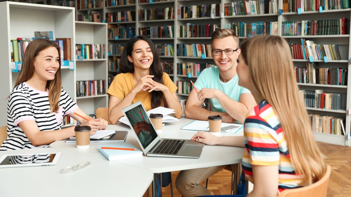 Vier junge Fachangestellte für Medien- und Informationsdienste sitzen in einer Bibliothek gemeinsam am runden Tisch und diskutieren über ein Projekt. Auf dem Tisch befinden sich Unterlagen, ein Laptop, ein Tablet und drei Kaffeebecher.
