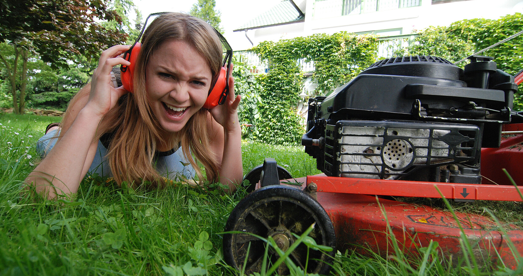 Mädchen mit Ohrenschützern liegt auf einer Wiese neben einem Rasenmäher