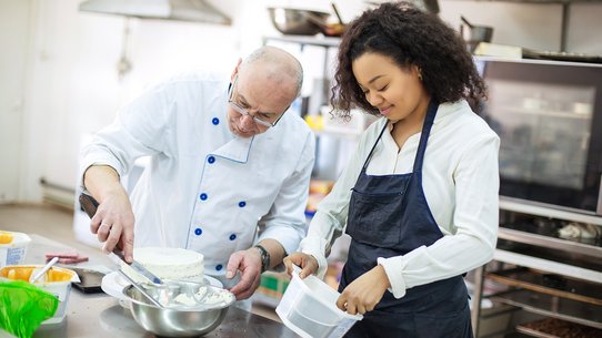 Auszubildende beim Kuchenbacken, sie rührt Teig an, der Konditor zeigt ihr, wie sie es machen soll