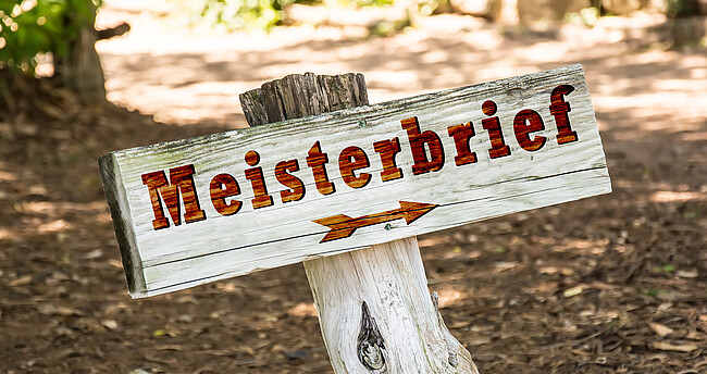 Holzschild mit brauner Beschriftung "Meisterbrief"; darunter zeigt ein Pfeil nach rechts