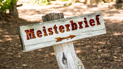 Holzschild mit brauner Beschriftung "Meisterbrief"; darunter zeigt ein Pfeil nach rechts