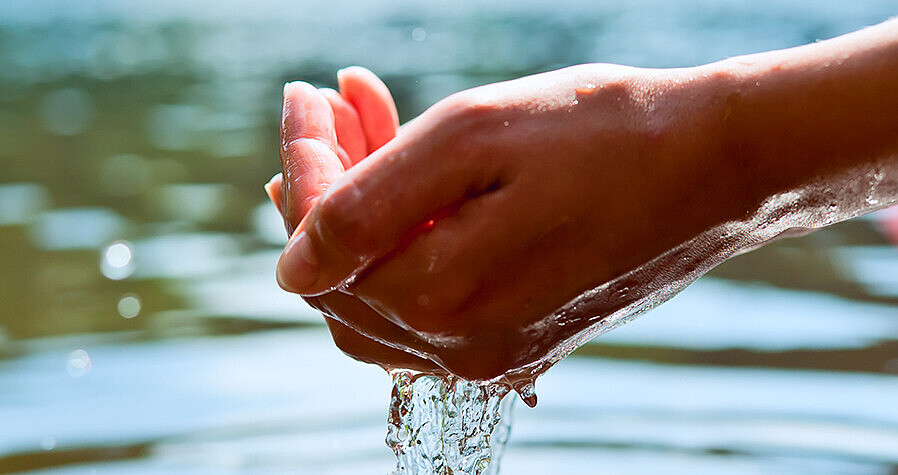Das Bild zeigt eine Hand, die Wasser aus einem See schöpft. 