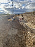 Ein breiter und tiefer Graben, der nach oben breiter wird. In der Sohle des Grabens ist ein Kipper-LKW zu sehen. Ein Bagger steht auf einer Rampe auf ungefähr halber Höhe des Grabens und schaufelt Erde. 
