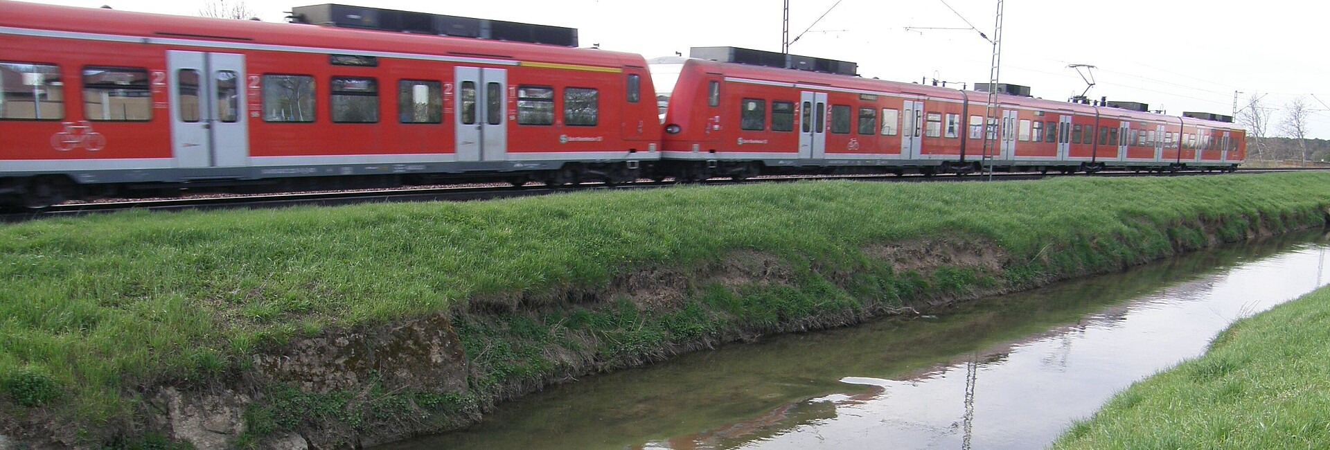 Auf dem Bild sieht man im Vordergrund den monotonen Gewässerlauf des Leimbachs und im Hintergrund die unmittelbar angrenzende parallel verlaufende Bahntrasse auf der gerade ein Zug fährt.