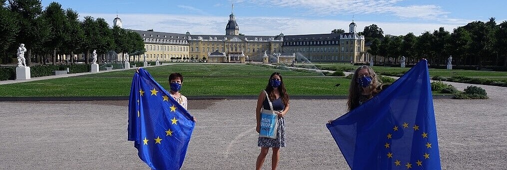 Im Vordergrund drei Frauen, alle tragen Mundschutzmasken mit der EU Flagge bedruckt. Zwei von Ihnen halten EU Flaggen vor sich, die Frau in der Mitte trägt eine Tasche mit dem Aufdruck „Mein Europa ist Zukunft“. Im Hintergrund ist unter blauem Himmel das Karlsruher Schloss zu sehen. 