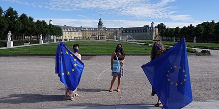 Im Vordergrund drei Frauen, alle tragen Mundschutzmasken mit der EU Flagge bedruckt. Zwei von Ihnen halten EU Flaggen vor sich, die Frau in der Mitte trägt eine Tasche mit dem Aufdruck „Mein Europa ist Zukunft“. Im Hintergrund ist unter blauem Himmel das Karlsruher Schloss zu sehen. 