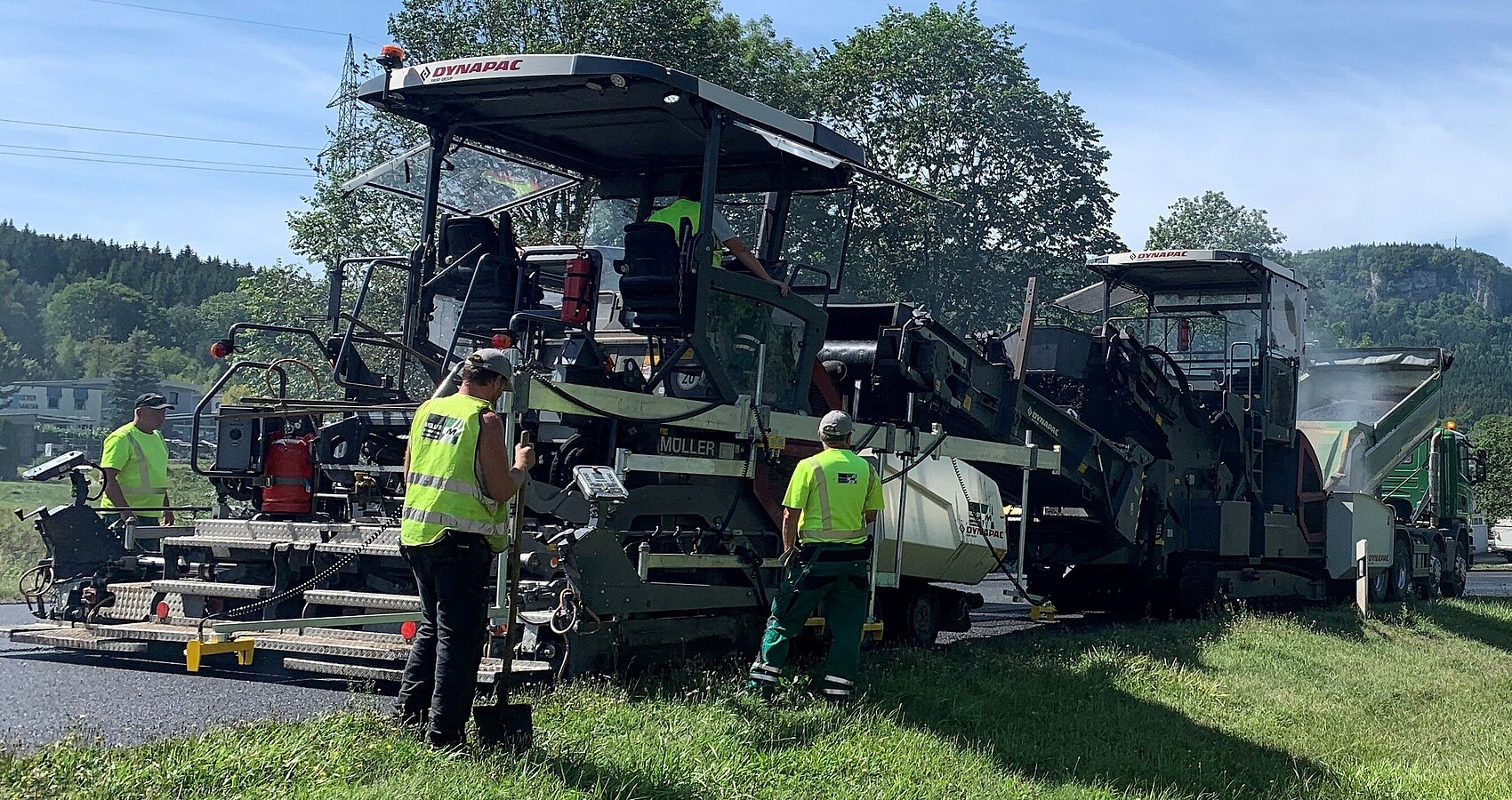 Man sieht ein großes Baustellenfahrzeug bei Asphaltierarbeiten und vier Personen in gelben Warnwesten