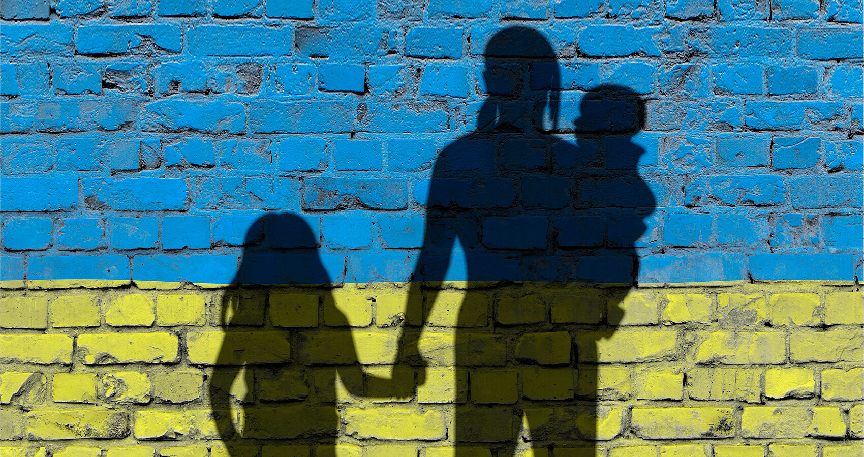 Bild zeigt Schattenumriss einer Frau mit einem Mädchen an der Hand und einem Kleinkind auf dem Arm vor einer Ziegelwand in den Farben der ukrainischen Flagge