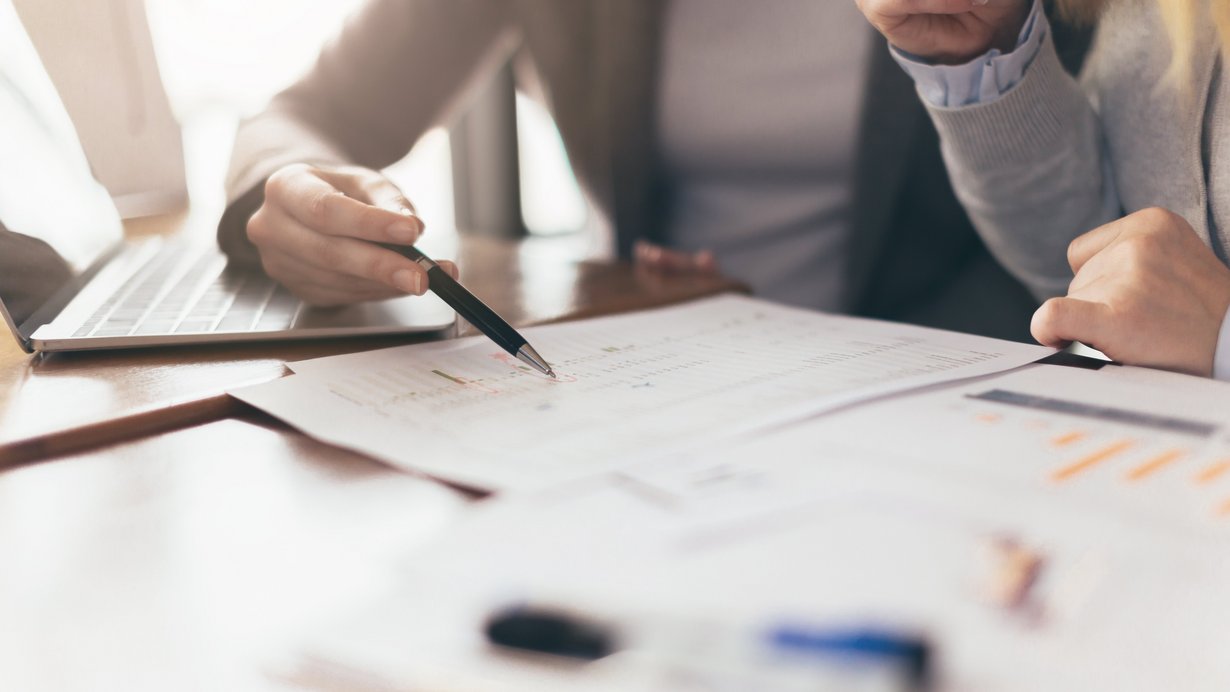 Zwei Kolleginnen arbeiten am Schreibtisch am Laptop und an Unterlagen, wie z.B. einer Statistik