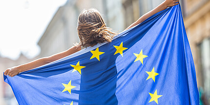 Junges Mädchen winkt mit der Flagge der Europäischen Union im Rücken in der Stadt.
