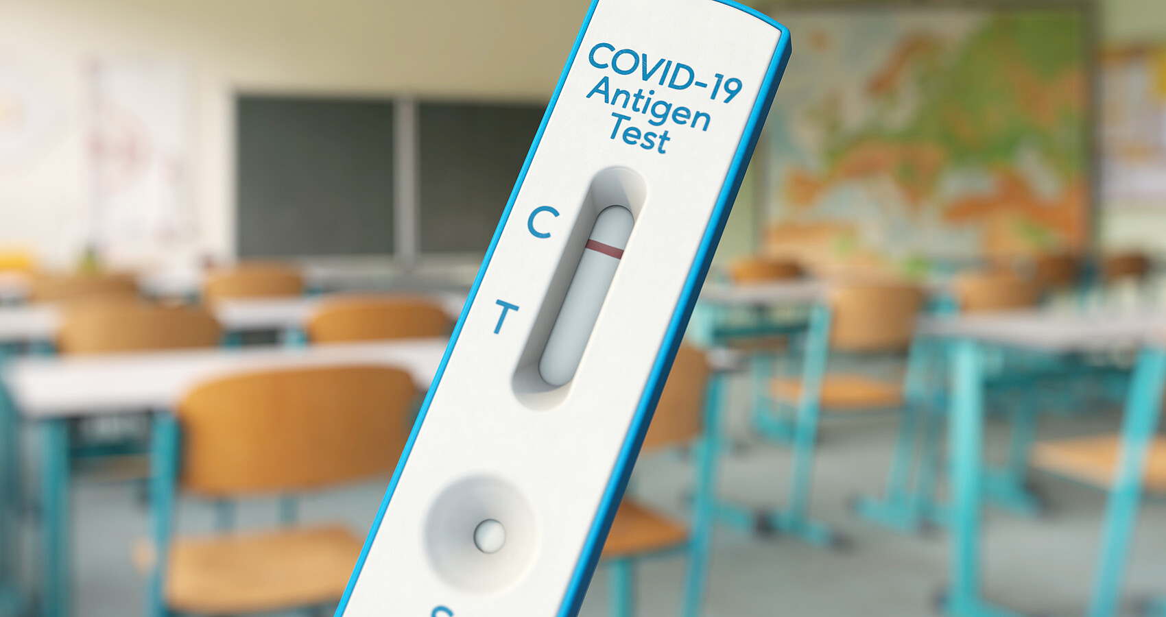 Corona-Test im Vordergrund des Bildes; im Hintergrund ist ein leeres Klassenzimmer zu sehen