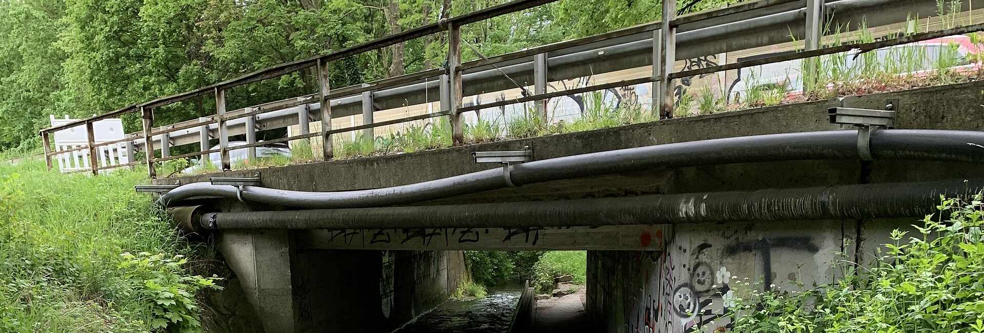 Eine Brücke von unten. Sie spannt ungefähr 10 Meter weit über einen kleinen Bach, an der Unterseite laufen Rohre.
