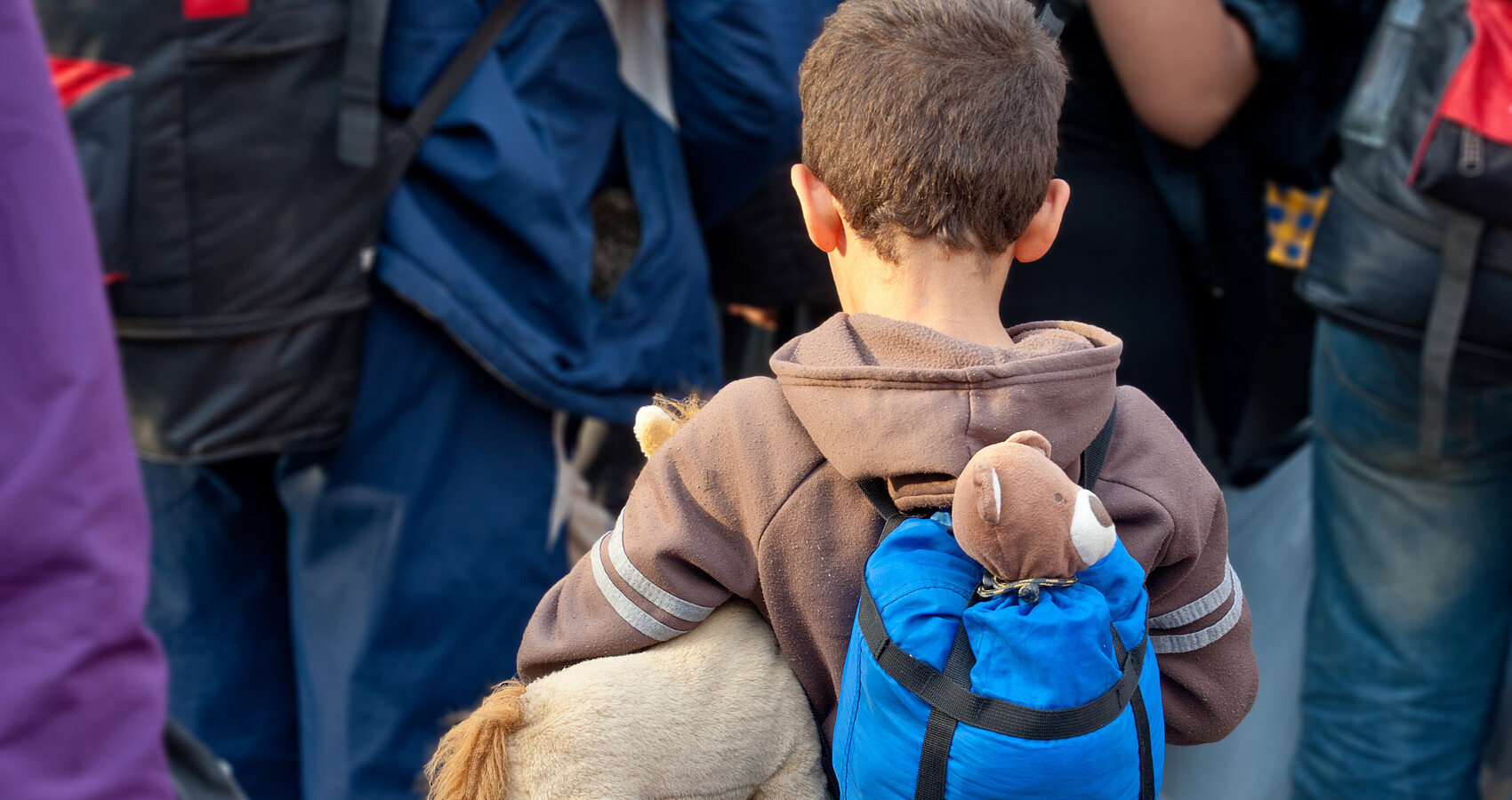 Bild zeigt Flüchtlinge, vorne ein Junge mit Rucksack, unter dem Arm ein Stofftier