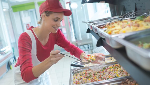 Fachpraktikerin der Hauswirtschaft bei der Essensausgabe, sie steht vor mehreren Behältnissen mit Speisen