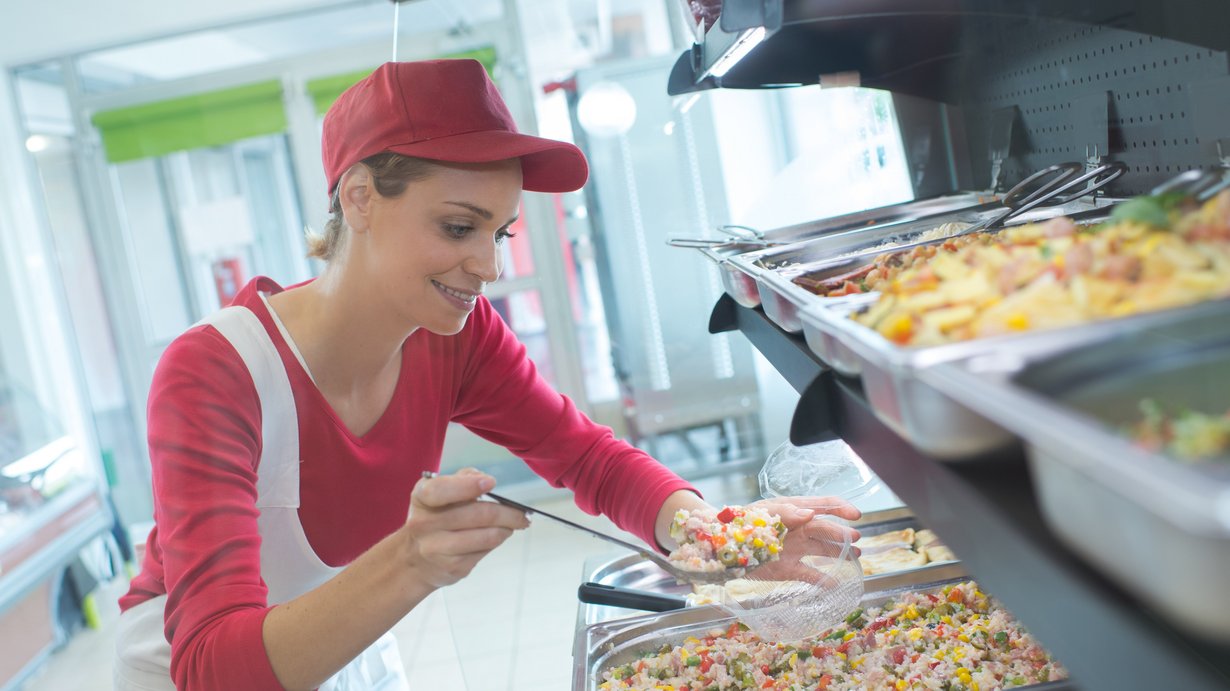 Fachpraktikerin der Hauswirtschaft bei der Essensausgabe, sie steht vor mehreren Behältnissen mit Speisen