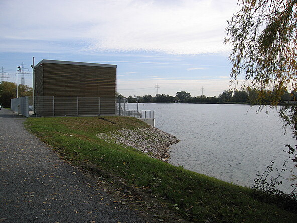 Außenansicht des Pumpwerks Freyersee mit Blick aus westlicher Richtung. Zu sehen ist das Betriebsgebäude, welches zur Schonung des Landschaftsbildes mit einer grünen Holzverkleidung versehen ist. Auf der rechten Bildseite ist zudem der Einlaufbereich des Pumpwerks sowie ein Teil des Freyersees zu sehen.“