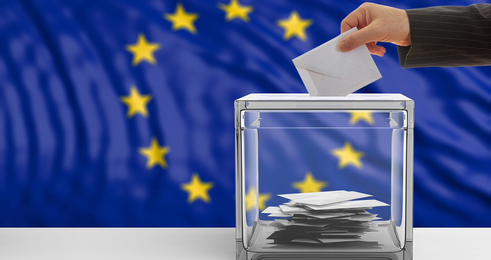 Im Hintergrund blaue EU Flagge mit den 12 gelben Sternen, im Vordergrund steht eine durchsichtige Wahlurne, in die ein verschlossener Wahlumschlag eingeworfen wird.
