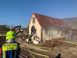 2025-02-04_UEbung_FFW_THW_Verschoenerungsverein.jpeg Ein Feuerwehrmann von hinten im Vordergrund. Im Hintergrund klettert ein Helfer des Technischen Hilfswerks über eine Leiter in den ersten Stock eines Gebäudes, aus dem Rauch kommt.