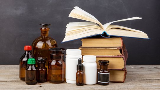 Bild zeigt Lehrbücher und Apothekenflaschen, die im Hörsaal vor einer Tafel auf dem Lehrpult stehen