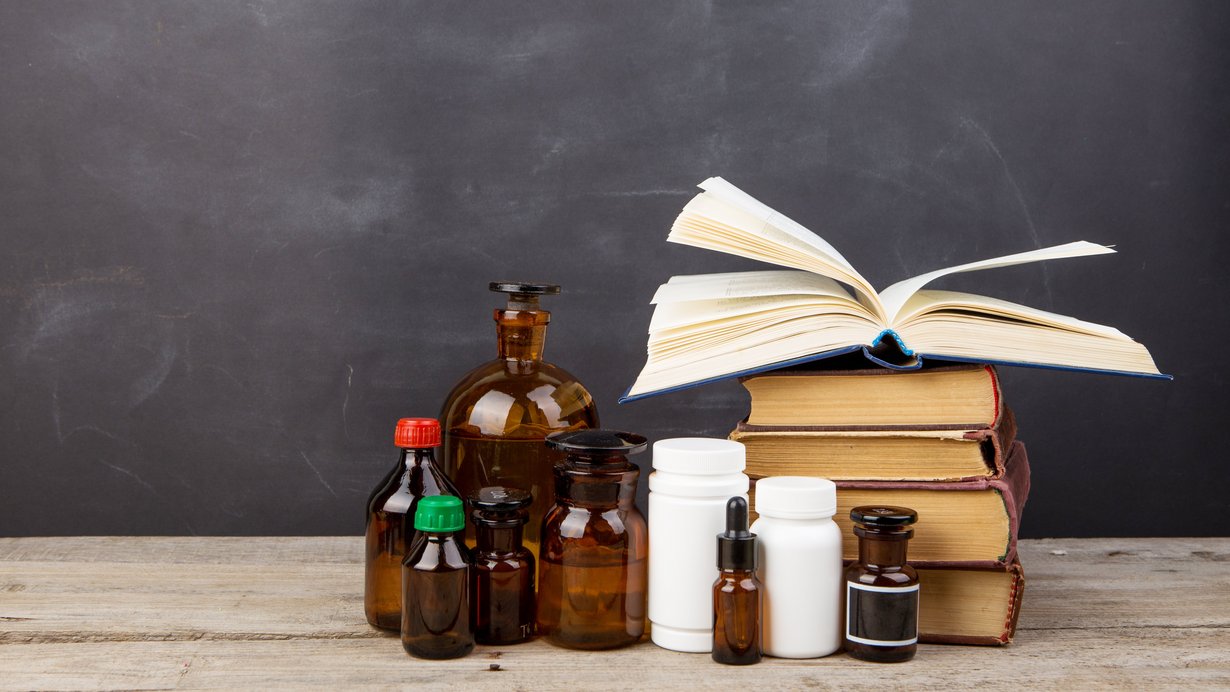 Bild zeigt Lehrbücher und Apothekenflaschen, die im Hörsaal vor einer Tafel auf dem Lehrpult stehen