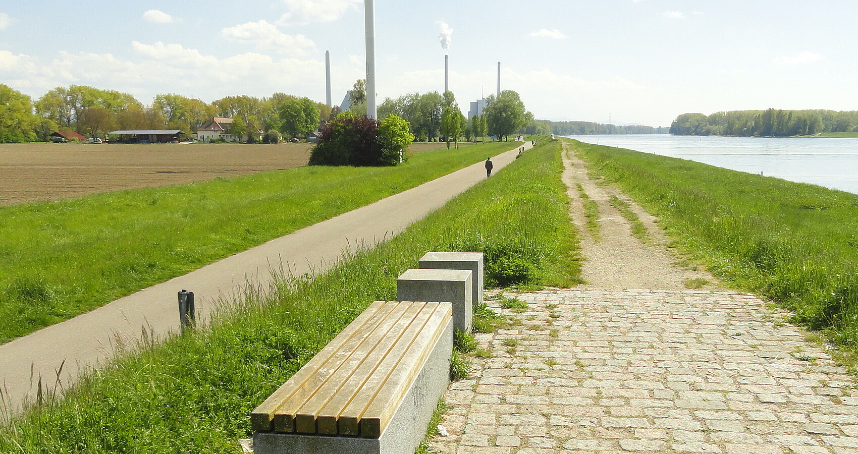 Auf dem Bild sieht man rechts den Rhein und links Wiesen und das Hofgut Maxau. Entlang des Rheins sieht man den unbefestigten Weg auf dem Rheindamm (rechts) sowie den befestigten Rad- und Fußweg am Dammfuß (links), mit Fußgänger. In Hintergrund befindet sich das Rheinhafen-Dampfkraftwerk, im Vordergrund einige Sitzgelegenheiten. Mittig, allerdings oben angeschnitten, befindet sich ein Windrad. 