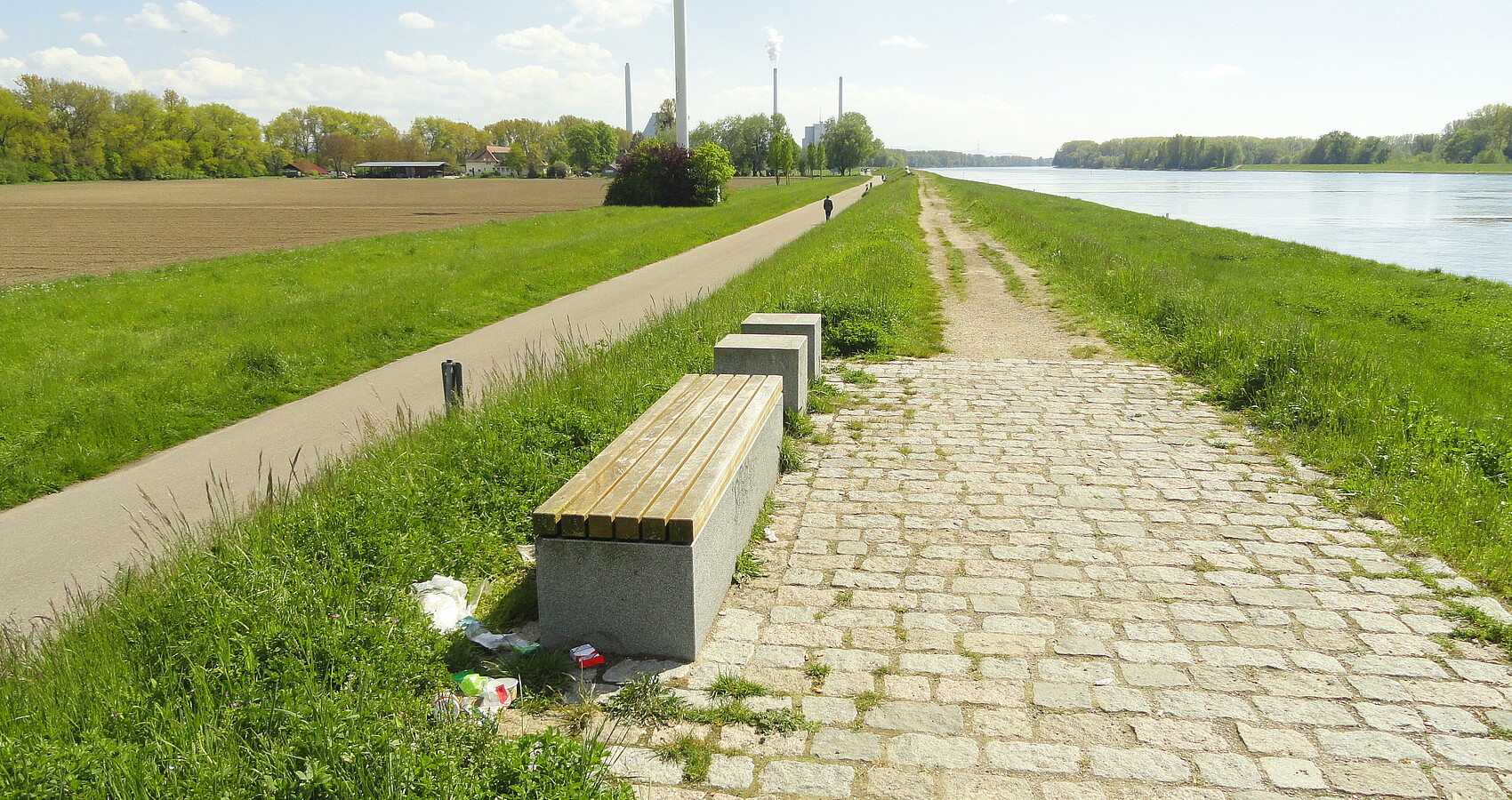 Auf dem Bild sieht man rechts den Rhein und links Wiesen und das Hofgut Maxau. Entlang des Rheins sieht man den unbefestigten Weg auf dem Rheindamm (rechts) sowie den befestigten Rad- und Fußweg am Dammfuß (links), mit Fußgänger. In Hintergrund befindet sich das Rheinhafen-Dampfkraftwerk, im Vordergrund einige Sitzgelegenheiten. Mittig, allerdings oben angeschnitten, befindet sich ein Windrad. 