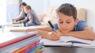 Ein Kind sitzt an seinen Hausaufgaben, dahinter hilft eine Frau einem weiteren Kind bei den Aufgaben