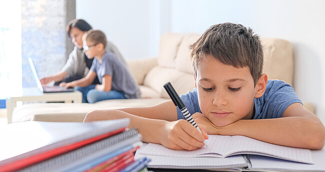 Ein Kind sitzt an seinen Hausaufgaben, dahinter hilft eine Frau einem weiteren Kind bei den Aufgaben