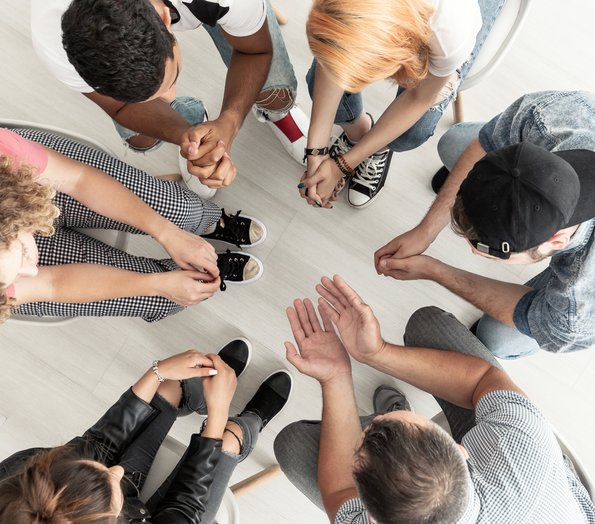 Fünf Jugendliche sitzen mit dem Lehrer in einem Sitzkreis, die Köpfe sind nach unten gesenkt und besprechen sich. 