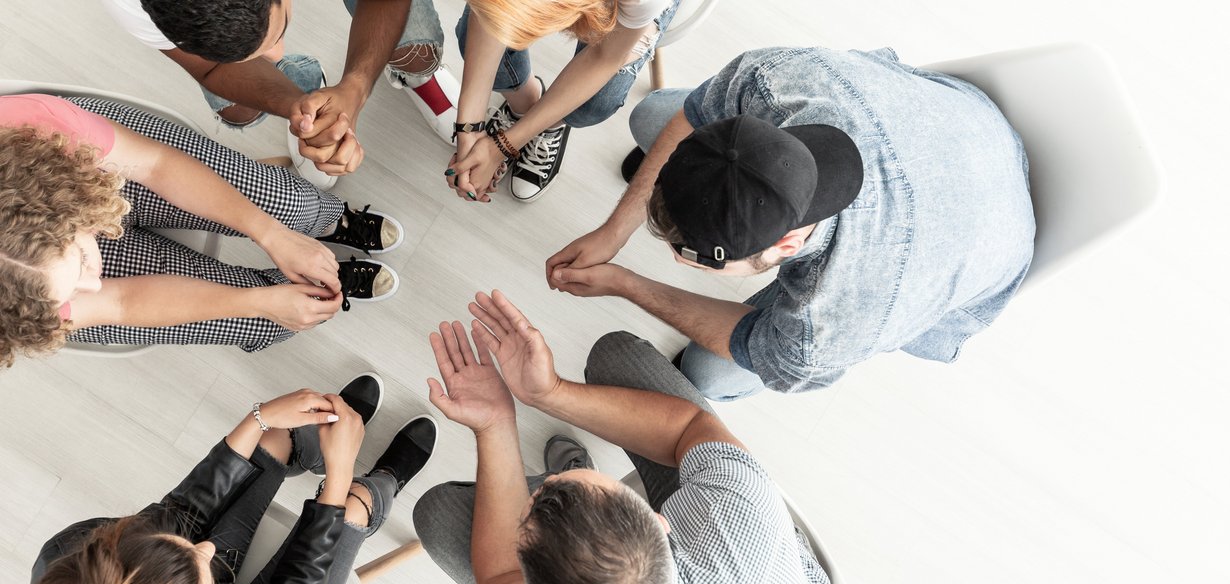 Fünf Jugendliche sitzen mit dem Lehrer in einem Sitzkreis, die Köpfe sind nach unten gesenkt und besprechen sich. 