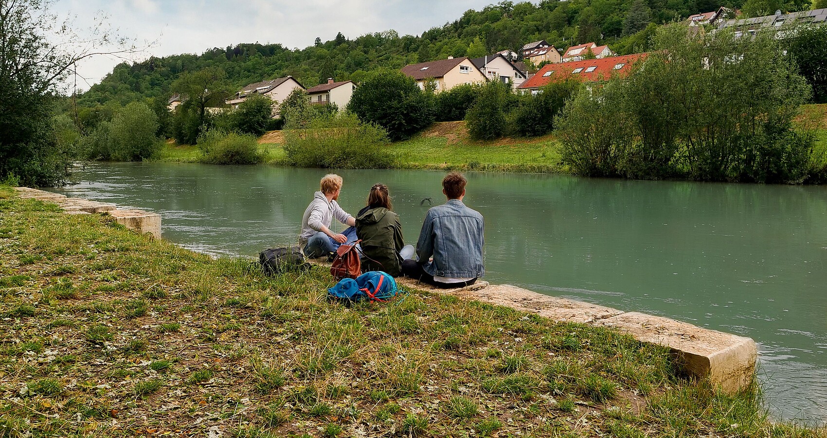 Drei junge Menschen sitzen am Ufer des revitalisierten Neckarabschnitt auf Höhe des Tübinger Freibads und schauen ins Wasser