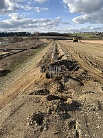 Ein Straßendamm aus Erde. Am Rand ragen kreisförmig angeordnete Baustahl-Stangen aus dem Boden. 