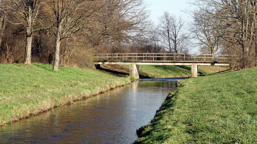 Ein gerade fließender Fluss fließt durch die Landschaft. Im Hintergrund sind eine Brücke und Bäume zu sehen. Die Ufer isnd gemäht und artenarm.