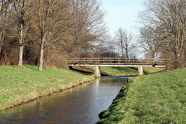 Ein gerade fließender Fluss fließt durch die Landschaft. Im Hintergrund sind eine Brücke und Bäume zu sehen. Die Ufer isnd gemäht und artenarm.