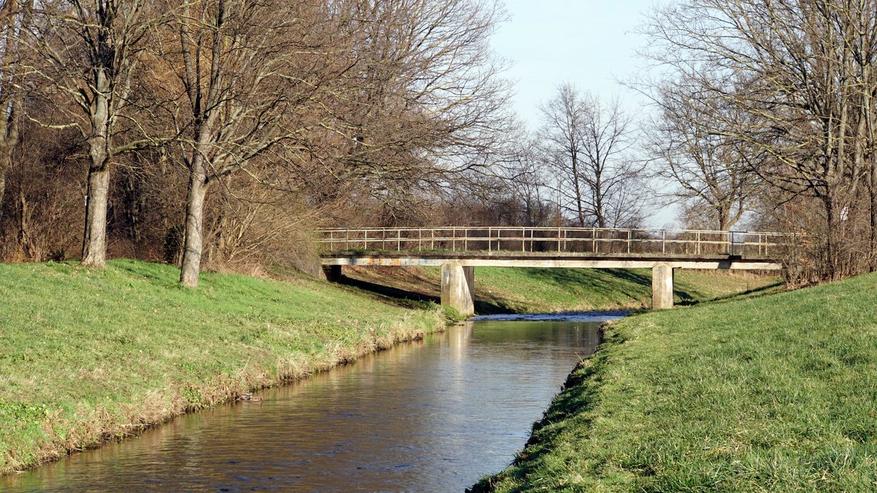 Ein gerade fließender Fluss fließt durch die Landschaft. Im Hintergrund sind eine Brücke und Bäume zu sehen. Die Ufer isnd gemäht und artenarm.