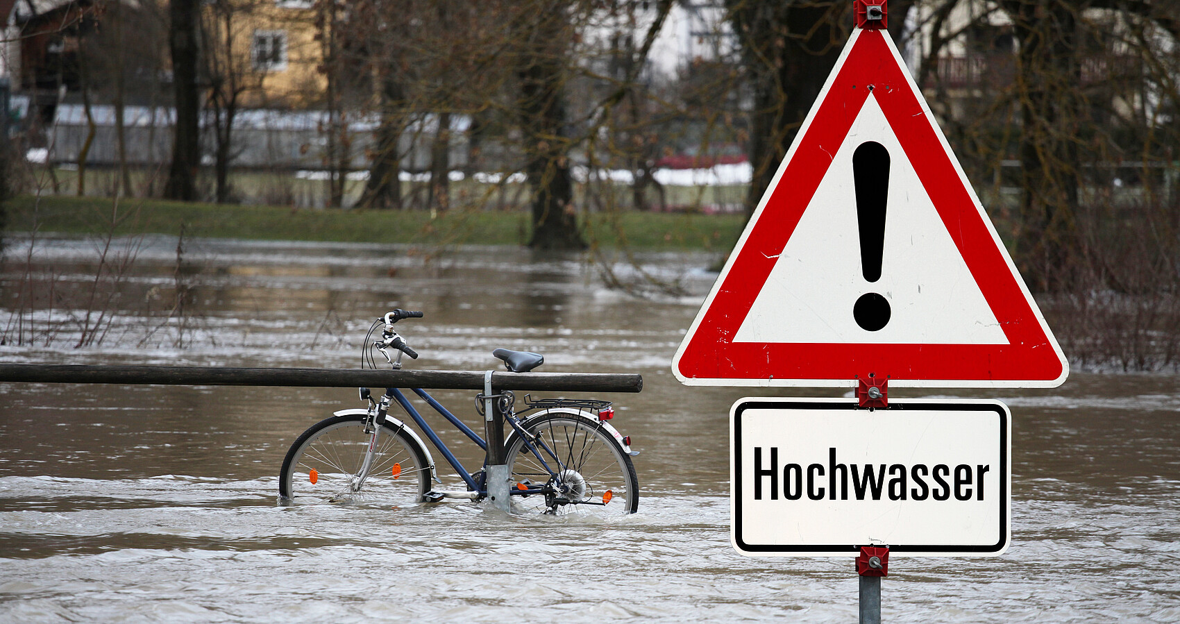 Gefahrenschild mit der Aufschrift "Hochwasser". Im Hintergrund sieht man Hochwasser mit einem Fahrrad