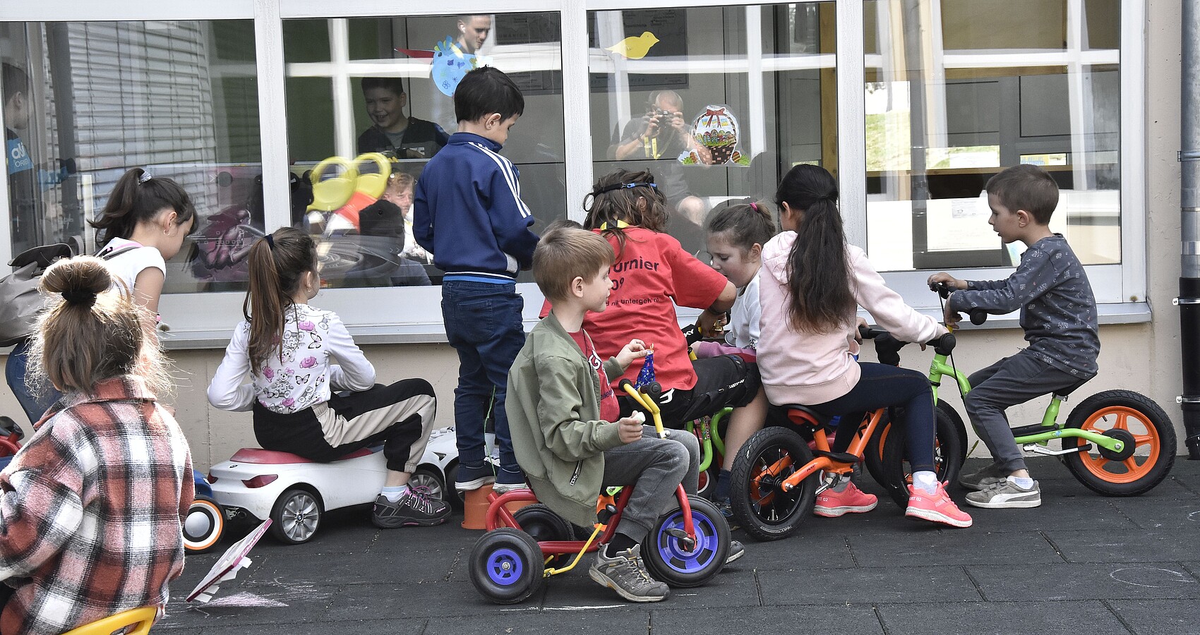 Das Bild zeigt eine Gruppe spielender Kindern auf Dreirädern, Laufrädern und Bobbycars vor einem großen Fenster eines Gebäudes