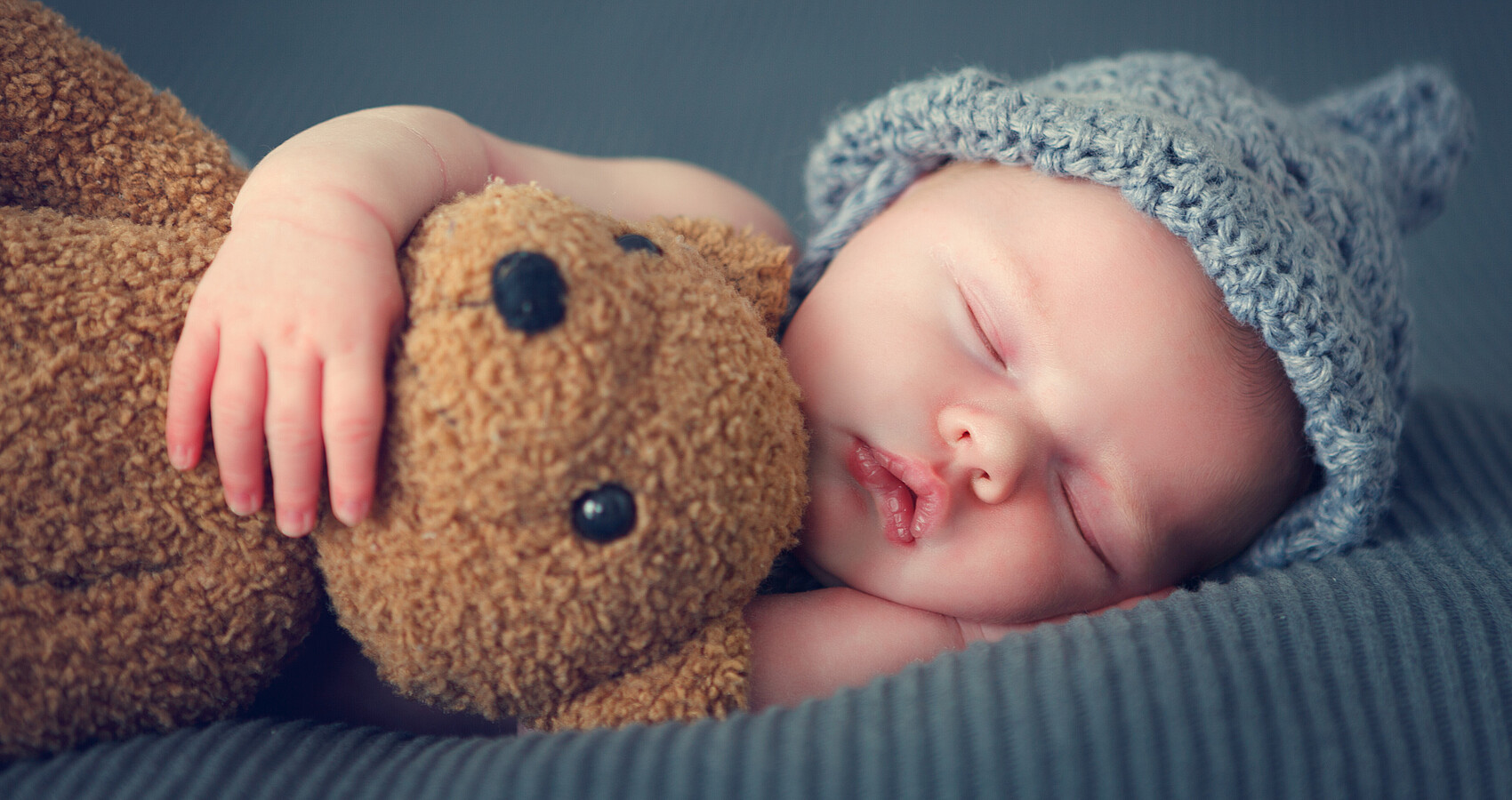 Man sieht ein schlafendes neugeborenes Baby mit einer blauen Mütze und einem Teddy im Arm