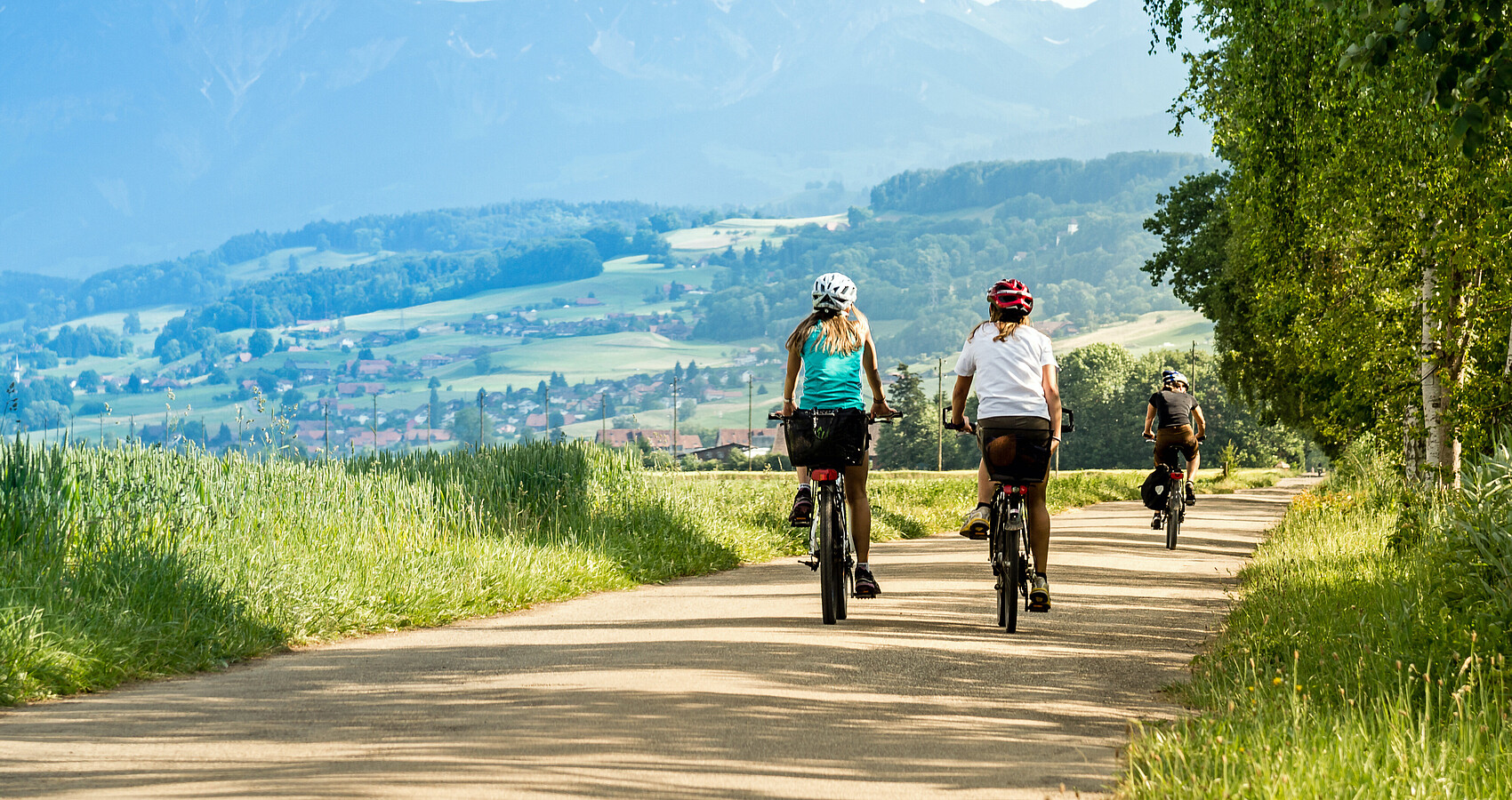 Zwei Radfahrerinnen sind auf einem Fahrradweg unterwegs, im Hintergrund Landschaft mit Bergen