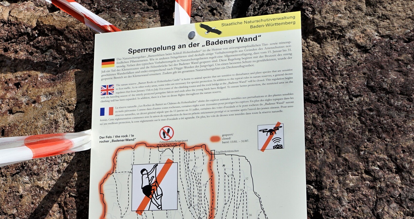 Informationstafel an einer Felswand mit rot-weißem Absperrband