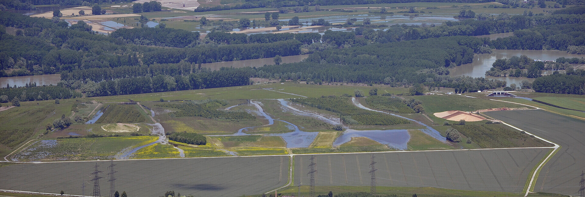 Luftbildaufnahme des Polder Rheinschanzinsel mit Blick in Richtung Norden. Zu sehen sind die mit Wasser gefüllten Gräben und Schluten im Bereich der Fläche für die partiellen Flutungen zur Auenrenaturierung. Im Hintergrund ist das überflutete Rheinvorland und auf der gegenüberliegenden Seite des Rheins der Polder Mechtersheim zu sehen.