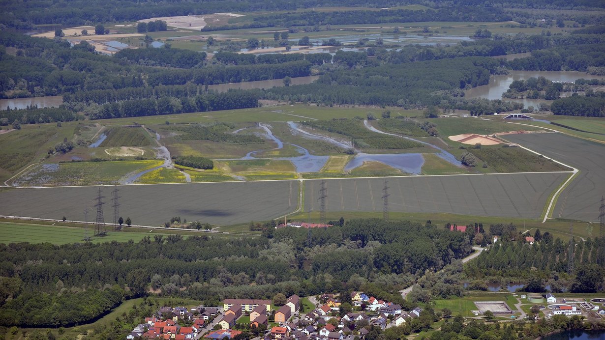 Luftbildaufnahme des Polder Rheinschanzinsel mit Blick in Richtung Norden. Zu sehen sind die mit Wasser gefüllten Gräben und Schluten im Bereich der Fläche für die partiellen Flutungen zur Auenrenaturierung. Im Hintergrund ist das überflutete Rheinvorland und auf der gegenüberliegenden Seite des Rheins der Polder Mechtersheim zu sehen.