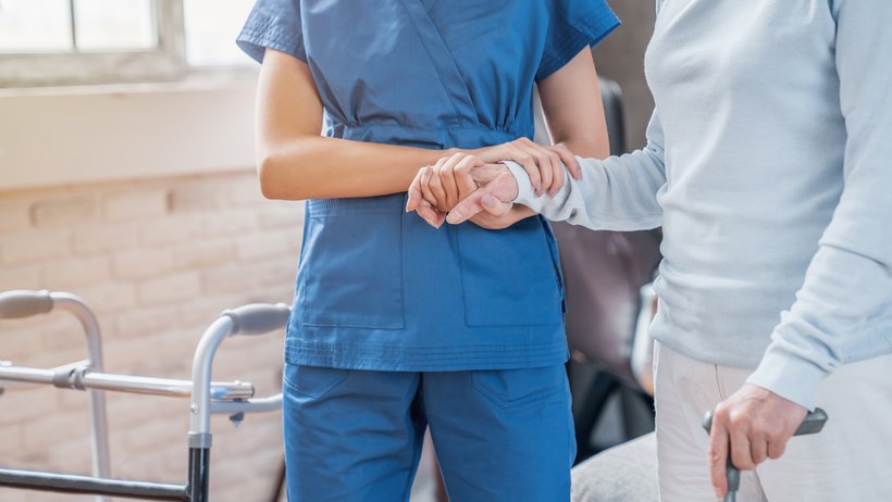 Bild zeigt eine Pflegekraft, die eine pflegebedürftige Person stütztend hält, die Person hält einen Gehstock in der Hand
