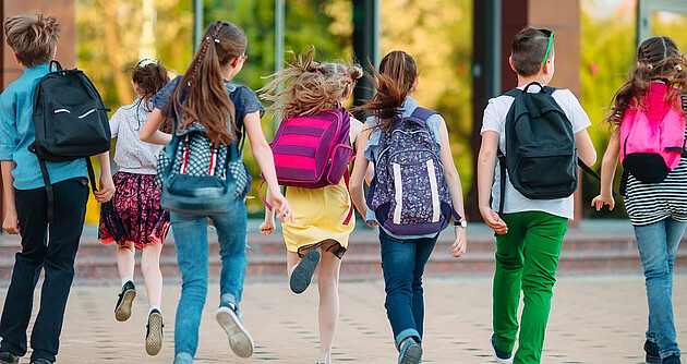 7 Schulkinder mit Rucksäcken rennen auf ein Schulgebäude zu