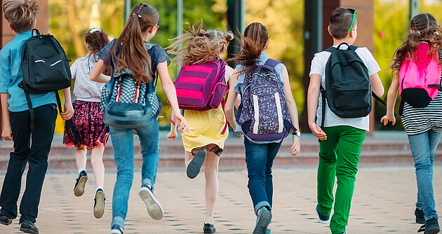 7 Schulkinder mit Rucksäcken rennen auf ein Schulgebäude zu