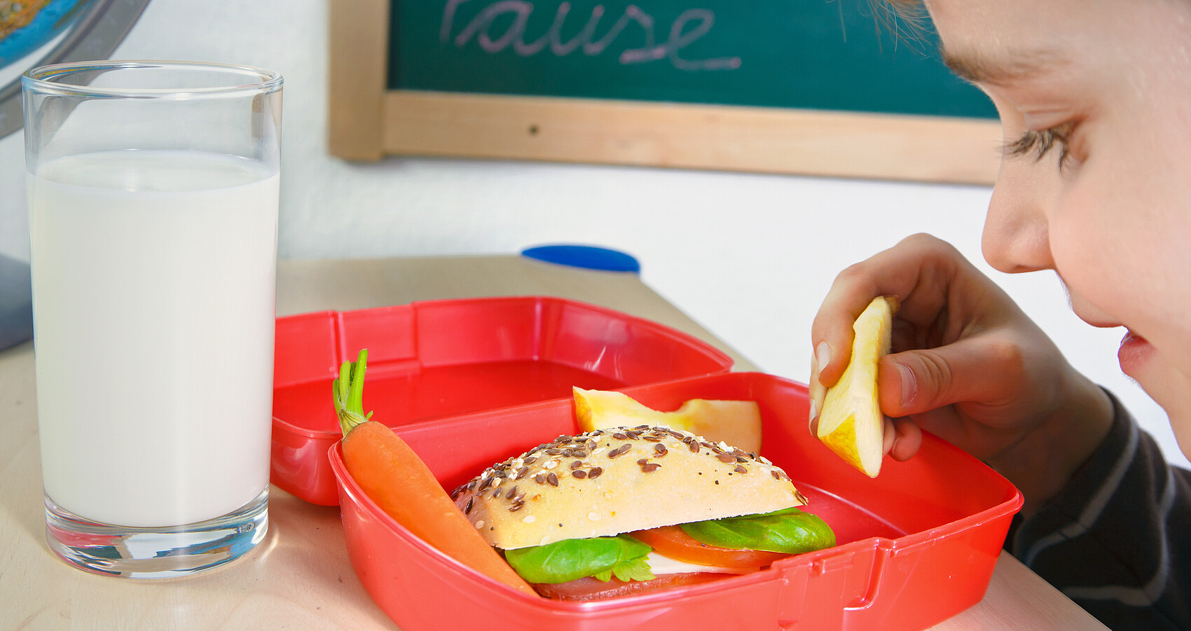 Kind blickt in Brotdose mit gesundem Frühstück aus Vollkornbrot, Äpfeln, Gemüse, davor ein Glas Milch