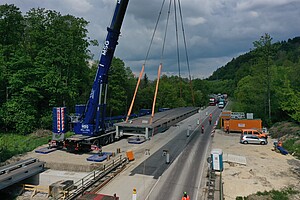 Ein Kran steht neben einer Straße. Er Hebt ein Brückenteil, das direkt neben der Straße liegt, auf circa einen Meter Höhe. Vor dem Kran befinden sich Brücken-Widerlager, auf denen bereits ein Brückenteil abgelegt ist.