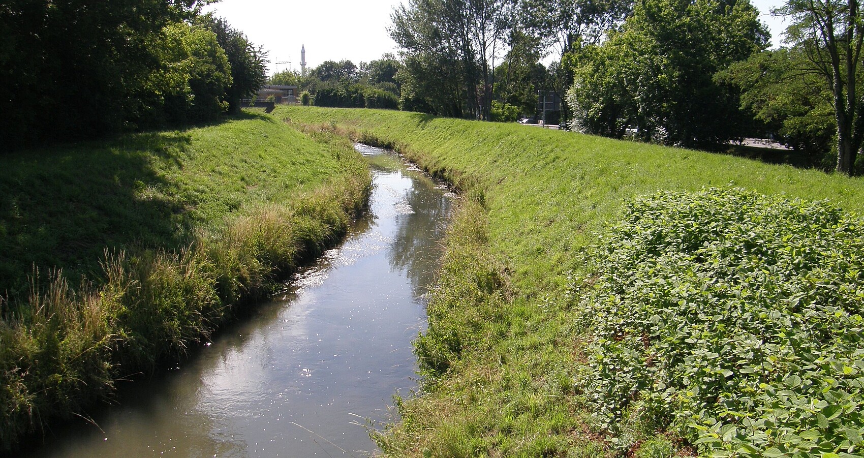 Auf dem Bild sieht man den monotonen und von seitlichen Dammstrukturen eingeengten Leimbach. Die Ufer und Dammbereiche sind mit Gras bewachsen. Auf beiden Seiten sieht man im Anschluss Bäume und Sträucher. Auf der rechten Seite erkennt man im Hintergrund eine Straße mit Straßenschild. In der Ferne sieht man eine Brücke, einen Turm sowie einen Kran.