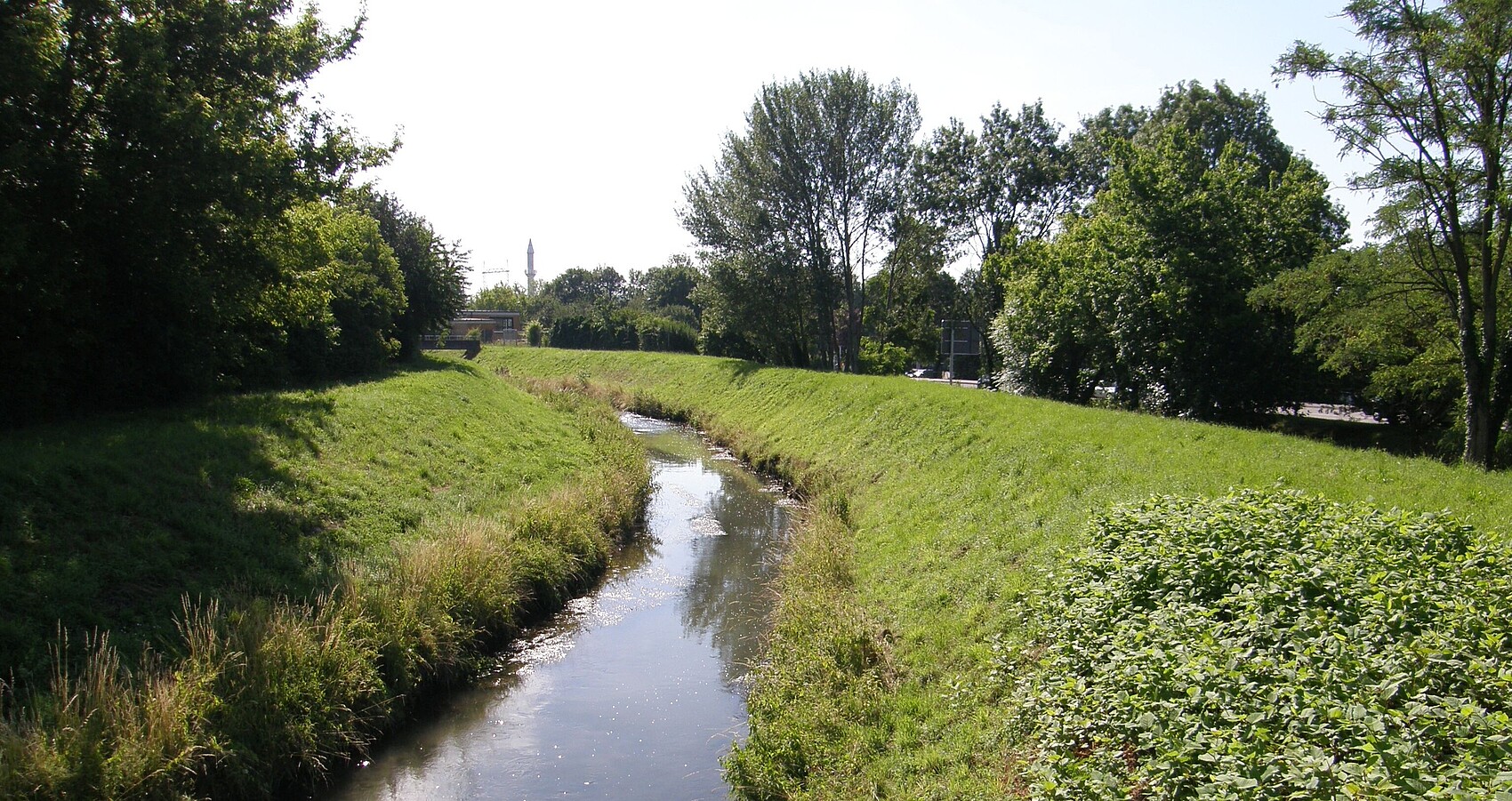 Auf dem Bild sieht man den monotonen und von seitlichen Dammstrukturen eingeengten Leimbach. Die Ufer und Dammbereiche sind mit Gras bewachsen. Auf beiden Seiten sieht man im Anschluss Bäume und Sträucher. Auf der rechten Seite erkennt man im Hintergrund eine Straße mit Straßenschild. In der Ferne sieht man eine Brücke, einen Turm sowie einen Kran.