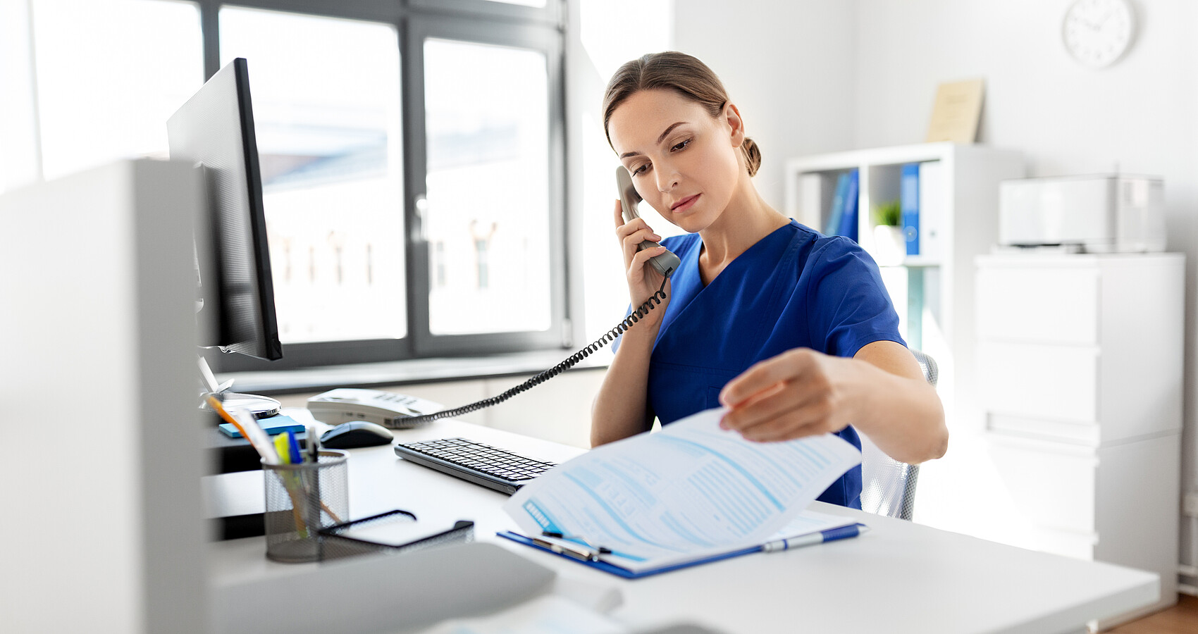 Eine Frau sitzt am Schreibtisch und blätter in Unterlagen während sie den Telefonhörer ans Ohr hält.